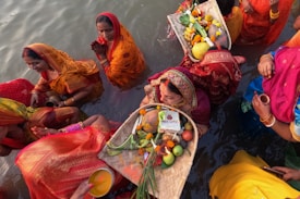 Several women dressed in vibrant traditional clothing are participating in a ritual by standing in the water. They are carrying baskets filled with fruits, offerings, and flowers. The scene is colorful, and the women appear focused and serene, engaging in a ceremonial activity.