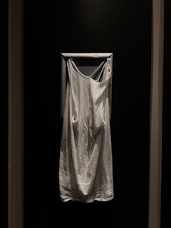 A white sleeveless garment hangs against a dark wall, framed by a white rectangular border. The fabric appears soft and loose-fitting.