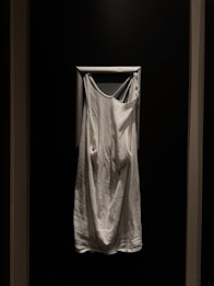 A white sleeveless garment hangs against a dark wall, framed by a white rectangular border. The fabric appears soft and loose-fitting.