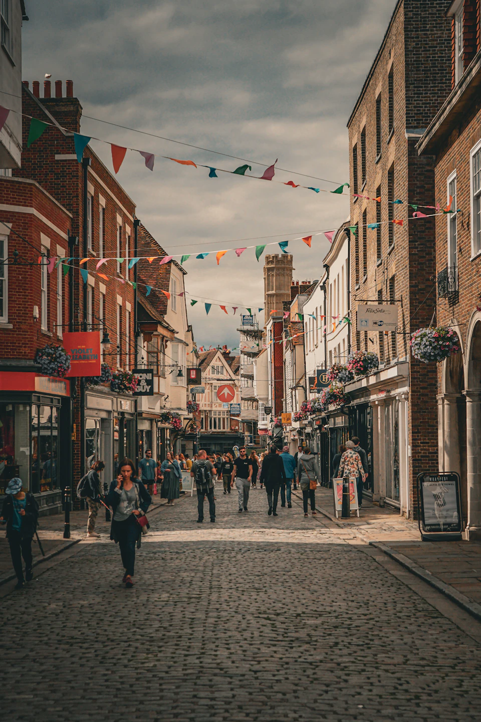 Bustling cobblestone street lined with brick buildings, colorful flags, and people walking along sidewalks