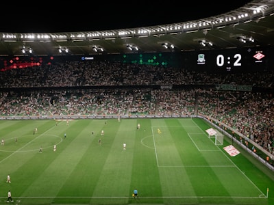 A soccer match is taking place in a large stadium filled with spectators. The scoreboard shows a score of 0:2, indicating that the away team is winning. Players are spread across the field, with a goalkeeper near one of the goals. The stadium is brightly lit and the stands are packed with fans.