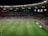 A soccer match is taking place in a large stadium filled with spectators. The scoreboard shows a score of 0:2, indicating that the away team is winning. Players are spread across the field, with a goalkeeper near one of the goals. The stadium is brightly lit and the stands are packed with fans.