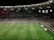 A soccer match is taking place in a large stadium filled with spectators. The scoreboard shows a score of 0:2, indicating that the away team is winning. Players are spread across the field, with a goalkeeper near one of the goals. The stadium is brightly lit and the stands are packed with fans.