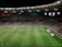 A soccer match is taking place in a large stadium filled with spectators. The scoreboard shows a score of 0:2, indicating that the away team is winning. Players are spread across the field, with a goalkeeper near one of the goals. The stadium is brightly lit and the stands are packed with fans.