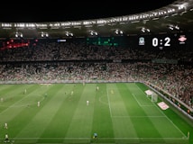 A soccer match is taking place in a large stadium filled with spectators. The scoreboard shows a score of 0:2, indicating that the away team is winning. Players are spread across the field, with a goalkeeper near one of the goals. The stadium is brightly lit and the stands are packed with fans.