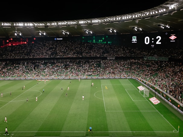 A soccer match is taking place in a large stadium filled with spectators. The scoreboard shows a score of 0:2, indicating that the away team is winning. Players are spread across the field, with a goalkeeper near one of the goals. The stadium is brightly lit and the stands are packed with fans.