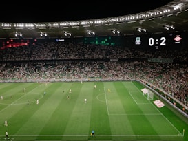 A soccer match is taking place in a large stadium filled with spectators. The scoreboard shows a score of 0:2, indicating that the away team is winning. Players are spread across the field, with a goalkeeper near one of the goals. The stadium is brightly lit and the stands are packed with fans.