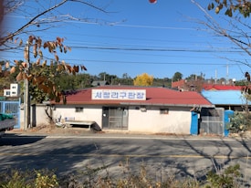 A small, single-story building with a red roof and a sign in Korean above the entrance. The structure is situated on a quiet road with an empty bench in front. There are some trees with autumn foliage in the foreground and background, and clear blue skies overhead.