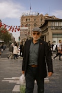 A bustling city street with pedestrians walking by, featuring a historic building in the background adorned with a sign reading 'Turkiye Bankasi.' Red flags with crescent moons and stars hang across the sky. Various people are walking, including a man in the foreground wearing a dark coat and holding a plastic bag.