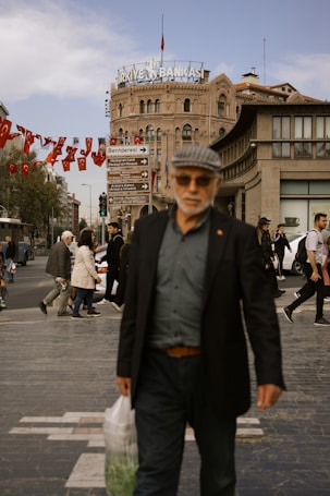 A bustling city street with pedestrians walking by, featuring a historic building in the background adorned with a sign reading 'Turkiye Bankasi.' Red flags with crescent moons and stars hang across the sky. Various people are walking, including a man in the foreground wearing a dark coat and holding a plastic bag.
