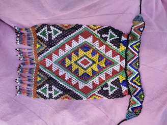 A colorful, intricately beaded textile featuring geometric patterns and fringed edges. The design includes a central diamond shape with concentric layers of colored shapes radiating outwards, utilizing vibrant hues such as blue, yellow, red, green, black, and white. The textile rests on a soft, light purple fabric background.