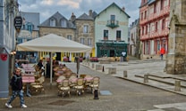 A picturesque European town square with charming historical architecture. Outdoor seating with checkered tablecloths is set up under a large cream-colored umbrella. The buildings surrounding the square feature a mix of beige stone and timber framing, painted in muted shades of green, red, and blue. People are casually walking or sitting at the café tables, contributing to a lively yet relaxed atmosphere.