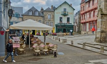 A picturesque European town square with charming historical architecture. Outdoor seating with checkered tablecloths is set up under a large cream-colored umbrella. The buildings surrounding the square feature a mix of beige stone and timber framing, painted in muted shades of green, red, and blue. People are casually walking or sitting at the caf&eacute; tables, contributing to a lively yet relaxed atmosphere.
