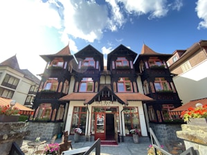 A picturesque villa with a classic European architectural style, featuring dark wooden beams and red-framed windows. The front garden is adorned with colorful flowers, and the building has a steep roof with pointed turrets. The stone facade and balconies with flower boxes add to its charm, set against a bright blue sky with scattered clouds.