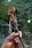 A hand is holding a violin neck with visible tuning pegs and strings. The violin has a striped ribbon attached. The background is blurred greenery, suggesting an outdoor setting.