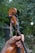 A hand is holding a violin neck with visible tuning pegs and strings. The violin has a striped ribbon attached. The background is blurred greenery, suggesting an outdoor setting.