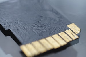 A close-up view of a black microchip or circuit board featuring intricate patterns and textures, with several gold connectors visible on one side.