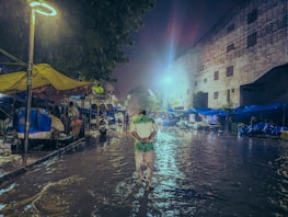 A person walks down a flooded street at night under a lit streetlamp. Market stalls with yellow and blue tarps line the sides, and water reflects the light, creating a moody atmosphere.