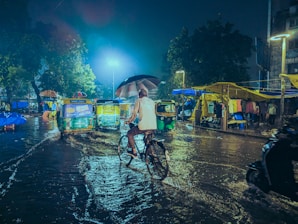 A person on a bicycle is seen riding through a flooded street at night, holding an umbrella. Rickshaws are parked on the wet road, and there's a market with yellow canopies along the right side. The scene is illuminated by streetlights and there are trees in the background.