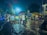 A person on a bicycle is seen riding through a flooded street at night, holding an umbrella. Rickshaws are parked on the wet road, and there's a market with yellow canopies along the right side. The scene is illuminated by streetlights and there are trees in the background.