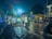 A person on a bicycle is seen riding through a flooded street at night, holding an umbrella. Rickshaws are parked on the wet road, and there's a market with yellow canopies along the right side. The scene is illuminated by streetlights and there are trees in the background.