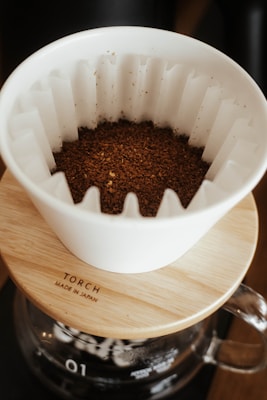A white ceramic coffee dripper sits atop a wooden lid with the word 'TORCH' and 'Made in Japan' engraved on it. The dripper contains freshly ground coffee. Below the dripper is a glass coffee pot.