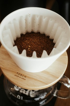 A white ceramic coffee dripper sits atop a wooden lid with the word 'TORCH' and 'Made in Japan' engraved on it. The dripper contains freshly ground coffee. Below the dripper is a glass coffee pot.