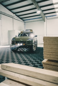 A rugged SUV with off-road modifications is parked inside a spacious industrial workshop. The vehicle has large tires and additional lighting mounted on the roof. The floor has a patterned surface, and wooden planks are stacked to one side.
