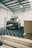 A rugged SUV with off-road modifications is parked inside a spacious industrial workshop. The vehicle has large tires and additional lighting mounted on the roof. The floor has a patterned surface, and wooden planks are stacked to one side.