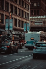 Vehicles, including a bus and a fire truck, are moving through a busy urban street lined with tall buildings and illuminated signs. The scene is captured under subdued lighting, suggesting an early morning or late afternoon setting.