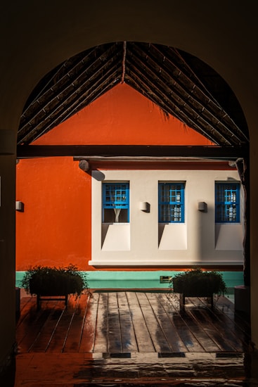 A vibrant courtyard scene with a striking contrast of colors, featuring a bright orange wall with two windows framed in vivid blue. The space is partially shaded by a sloped roof structure made of rustic wooden beams. Below the windows, a smooth wooden deck is present with two potted plants placed symmetrically on the sides, and a light teal trim runs along the base of the wall.