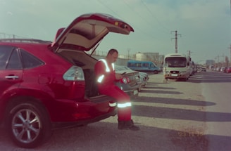 A person dressed in reflective red clothing is sitting on the open tailgate of a red car, parked alongside other vehicles on a gravel road. The car's trunk is open, and several other cars and a bus can be seen parked in a line. The sky is cloudy, and there are power lines overhead.