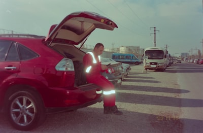 A person dressed in reflective red clothing is sitting on the open tailgate of a red car, parked alongside other vehicles on a gravel road. The car's trunk is open, and several other cars and a bus can be seen parked in a line. The sky is cloudy, and there are power lines overhead.