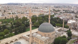 Aerial view of a large mosque with a prominent dome and four tall minarets, surrounded by lush greenery and an urban landscape. The mosque appears to be a central historical architectural structure within a sprawling city.
