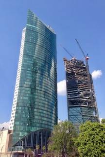 Two tall skyscrapers, one fully constructed and the other under construction, with a blue sky in the background. The completed building has a curved glass facade with greenish-tinted windows, while the building under construction features exposed structural elements and cranes. Green foliage is visible at the bottom of the image.