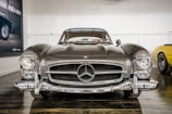 A classic Mercedes-Benz car is prominently displayed, showcasing its sleek design and vintage appeal. The front view highlights the iconic grille and circular headlights. The car is parked indoors with reflective flooring and a minimalistic backdrop. Another vintage car is partially visible to the right.