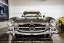 A classic Mercedes-Benz car is prominently displayed, showcasing its sleek design and vintage appeal. The front view highlights the iconic grille and circular headlights. The car is parked indoors with reflective flooring and a minimalistic backdrop. Another vintage car is partially visible to the right.