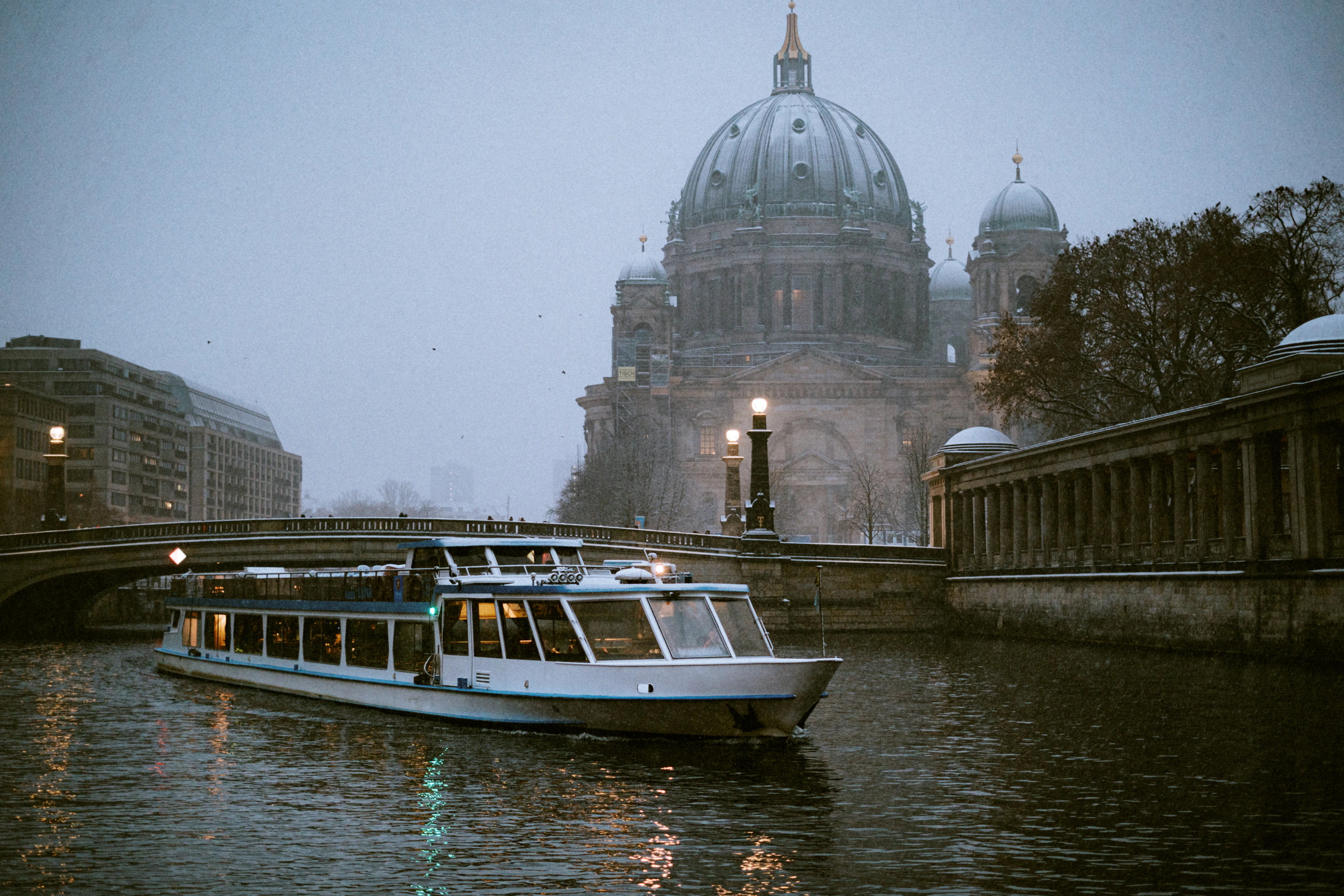 A glass-roofed sightseeing boat on the Spree passing beneath the domes of Berlin Cathedral on a misty winter morning.
