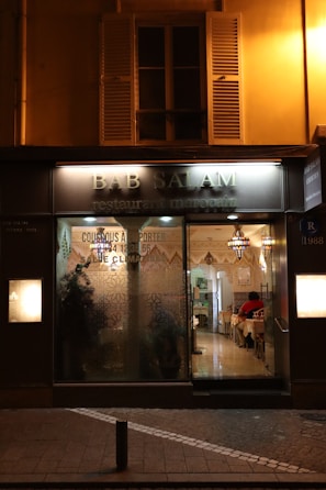Exterior view of a Moroccan restaurant named Bab Salam with warm lighting illuminating the entrance. The glass facade features decorative designs, offering a glimpse into a cozy interior where patrons sit at tables covered with white tablecloths. Traditional Moroccan lamps hang from the ceiling, enhancing the inviting atmosphere.