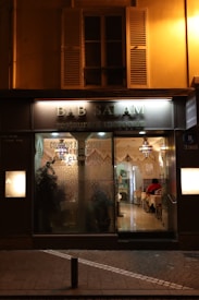 Exterior view of a Moroccan restaurant named Bab Salam with warm lighting illuminating the entrance. The glass facade features decorative designs, offering a glimpse into a cozy interior where patrons sit at tables covered with white tablecloths. Traditional Moroccan lamps hang from the ceiling, enhancing the inviting atmosphere.