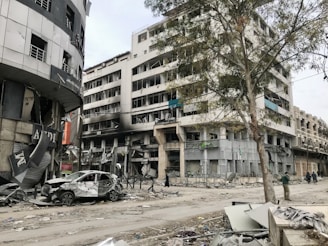 A scene of urban devastation featuring a severely damaged multi-story building with broken windows and a charred facade. The surrounding area is littered with debris and a wrecked car is parked among the rubble. A large tree stands near the building, and a few people are seen walking along the street.