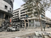 A scene of urban devastation featuring a severely damaged multi-story building with broken windows and a charred facade. The surrounding area is littered with debris and a wrecked car is parked among the rubble. A large tree stands near the building, and a few people are seen walking along the street.