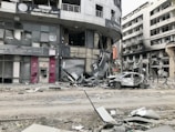 A city street scene showing significant damage to buildings and vehicles. Storefronts are heavily damaged, with debris scattered around. A car with extensive damage is parked on the street next to debris and rubble. The surrounding multi-story buildings have broken windows and show signs of destruction.