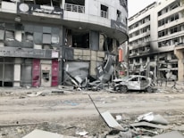 A city street scene showing significant damage to buildings and vehicles. Storefronts are heavily damaged, with debris scattered around. A car with extensive damage is parked on the street next to debris and rubble. The surrounding multi-story buildings have broken windows and show signs of destruction.