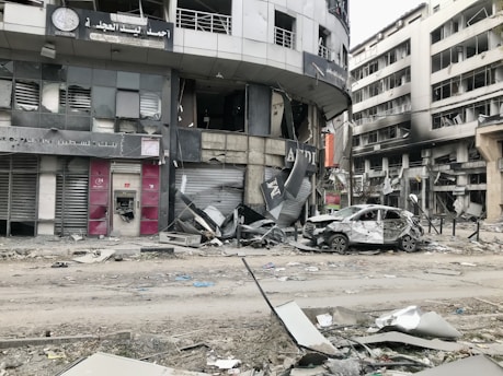 A city street scene showing significant damage to buildings and vehicles. Storefronts are heavily damaged, with debris scattered around. A car with extensive damage is parked on the street next to debris and rubble. The surrounding multi-story buildings have broken windows and show signs of destruction.