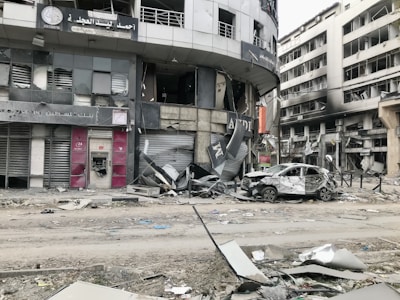 A city street scene showing significant damage to buildings and vehicles. Storefronts are heavily damaged, with debris scattered around. A car with extensive damage is parked on the street next to debris and rubble. The surrounding multi-story buildings have broken windows and show signs of destruction.