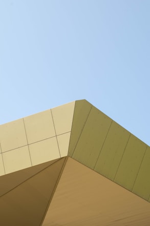 Angular architecture featuring clean lines and sharp edges with a minimalistic style against a clear blue sky. The building appears to have a modern design with sections in varying shades of beige and light green.