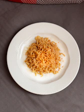 A white plate containing a serving of cooked instant noodles, placed on a textured gray surface. The noodles have a light golden color and appear to be plain without any garnishes.