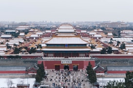A vast and intricate complex featuring traditional East Asian architecture with yellow-tiled roofs covered lightly with snow. Numerous buildings are visible, arranged in a symmetrical layout with courtyards and pathways. People are gathered near the entrance, and the surrounding landscape is wintry.