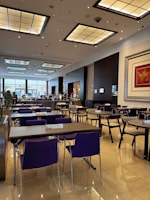 A spacious and modern dining area with neatly arranged tables and chairs. The room features a high ceiling with large, decorative light panels. The floor is polished with a reflective surface, and there's a large piece of artwork on the wall. Some people are visible in the background near the buffet area.